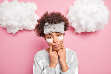 Young sad curly woman forces smile keeps fingers near corners of lips looks unhappily at camera wears sleepmask pajama has sleepy expression as awakes early in morning stands indoor feels upset