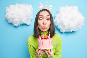 Surprised brunette Asian woman holds breath before blowing candles makes wish over birthday cake...
