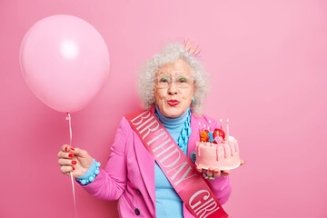 Fototapeta na wymiar Aged curly haired senior wrinkled woman keeps lips rounded holds delicious cake with burning candles inflated balloon celebrates 91st birthday wears festive clothes isolated over pink background
