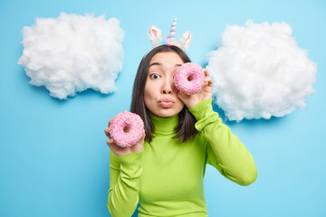 Portrait of attractive Asian woman covers eye with tasty doughnut keeps lips folded going to eat baked delicious sweet dessert wears unicorn headband green turtleneck isolated over blue wall