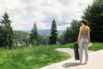 Fototapeta na wymiar Young fit woman with bottle of water and yoga mat looking at mountains. Outdoor workout, training