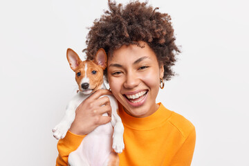 Portrait of good looking cheerful African American woman enjoys company of small pedigree dog wears...