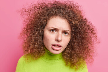 Headshot of indignant shocked young woman with curly hair looks unhappily at camera frowns face dressed in casual green turtleneck isolated over pink background. Human face expressions concept
