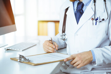 Friendly red-bearded doctor sitting and writing with clipboard in clinic, close-up