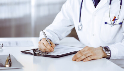 Unknown male doctor sitting and working with clipboard of medication history record in clinic at his working place, close-up. Young physician at work. Perfect medical service, medicine concept