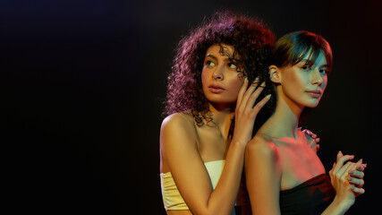 Two attractive half naked young brunette women with professional art makeup posing together in neon light isolated over black background