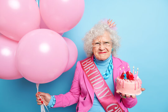 Portrait Of Senior Woman With Curly Grey Hair Has Dissatisfied Face Expression Wears Festive Clothes Celebrates Birthday Holds Tasty Cake Bunch Of Inflated Balloons Isolated Over Blue Background