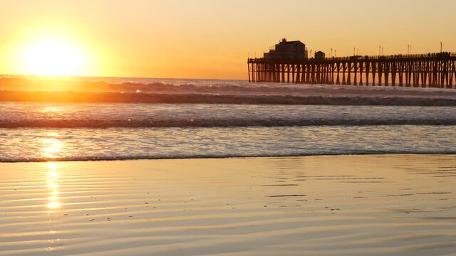 Wooden pier on piles, silhouette at sunset, California USA, Oceanside. Waterfront resort, pacific ocean tide, tropical beach. Summertime coastline vacations atmosphere. Sunny sea waves at sundown.