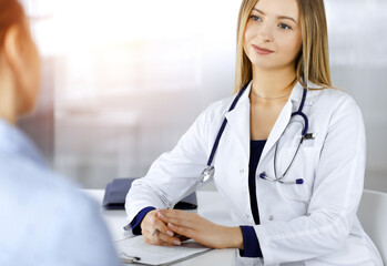 Young intelligent woman-doctor is listening to her patient's allergy's symptoms, while they are sitting together in the sunny cabinet in a clinic. Female physician, with a stethoscope, is writing at