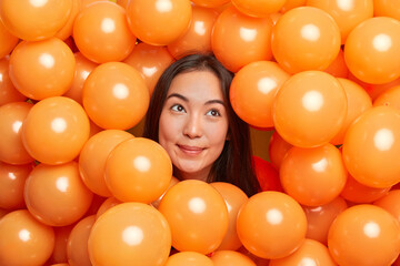 Dreamy brunette beautiful Asian woman concentrated above poses around inflated balloons thinks about holiday has satisfied expression decorates room for special occasion. Celebration concept