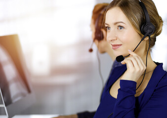 Two busineswomen have conversations with the clients by headsets, while sitting at the desk in a...