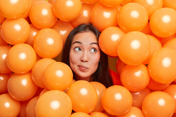 Fototapeta na wymiar Photo of cute thoughtful young Asian woman purses lips concentrated above with deep thoughts how to celebrate birthday sticks out head through small inflated orange balloons. Holidays concept