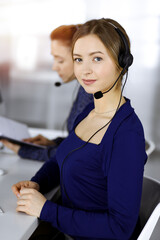 Two busineswomen have conversations with the clients by headsets, while sitting at the desk in a...