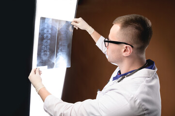 A young male doctor in glasses with a stethoscope around his neck holds an X-ray image of the spine...