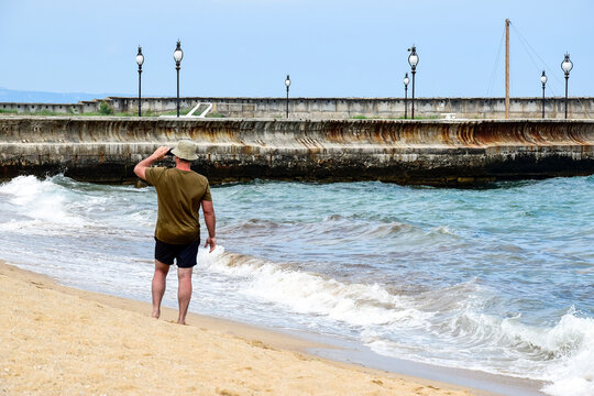 Man in T-shirt, shorts and panama hat stands on shore and takes pictures of stormy sea on smartphone. Back view. Big gray waves roll ashore, danger.
