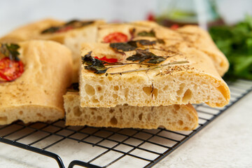 Slice of chopped focaccia, pizza, cut flat bread with vegetables on grid, tradition italian cuisine. Homemade artisan flatbread with tomatoes, parsley on white pastel table. Side view, close up