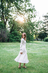 Naklejka premium Portrait of beautiful caucasian woman in long white retro dress walking in green summer blossom park on meadow. Rays of sunlight. Lady girl. Book cover