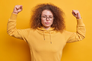 Serious beautiful European with curly hair looks confident at camera raises arms shows muscles...