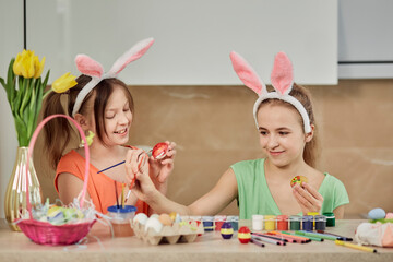 Two beautiful girls in bunny ears paint Easter eggs with paint and paint brush. Easter holiday.