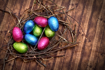 Foil wrapped colourful easter eggs in pink, green, blue and yelow in a natural nest made of sticks and twigs, against a multi grain brown wooden background.
