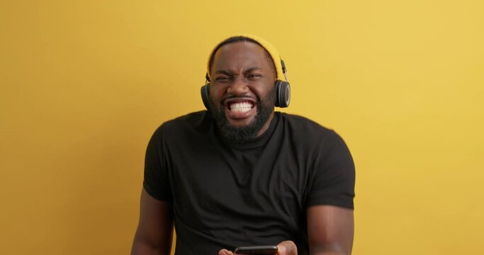 Handsome bearded black man chooses song from playlist holds modern smartphone listens music via stereo headphones wears casual black t shirt moves carefree against yellow background. Sound on