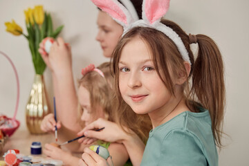 A cute girl paints an Easter egg and looks at the camera. Celebrating with the whole family. The traditional holiday is Easter.