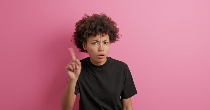 Displeased dark skinned woman shakes index fnger says no I dont agree with this prohibits bad offer looks gloomy dressedin casual black t shirt isolated over pink background. Hold on I forbid you