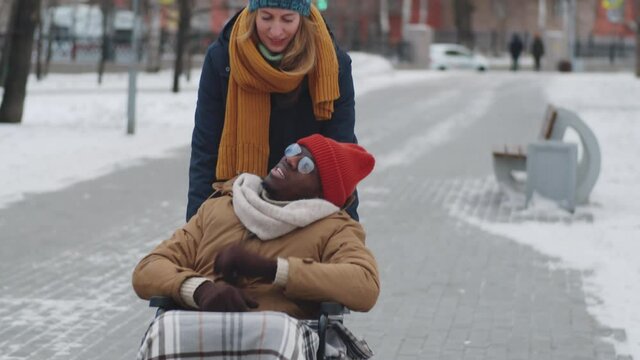 Young Caucasian woman pushing Afro-American man on wheelchair and talking with him during walk on winter day