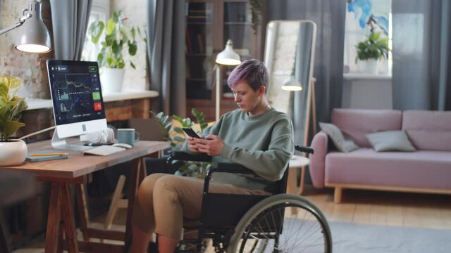 Young disabled woman on wheelchair typing on smartphone while having break during workday in home office