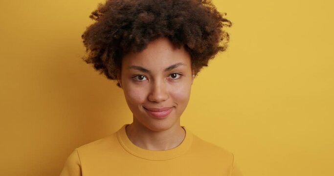 Lovely serious Afro American woman with dark curly hair looks directly at camera with determined assertive expression has little smile on face isolated over yellow background wears casual clothes