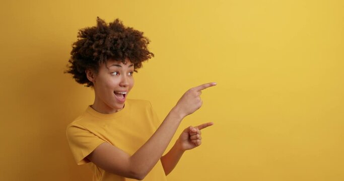 Positive curly haired woman smiles positively recommends using blank space indicates at upper right corner dressed in casual t shirt isolated over vivid yellow background shows direction or way