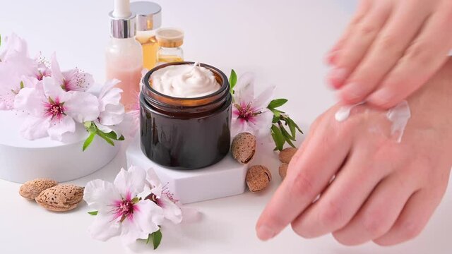 A woman smears her hands with cream. Hand care lotion with almond oil and vitamins. Body skin health and beauty. A bright background, jars with cosmetics and natural almond flowers. 