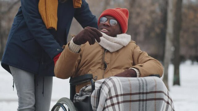 Tilt up shot of young Caucasian woman and cheerful Afro-American man on wheelchair chatting and laughing outdoors on street on winter day