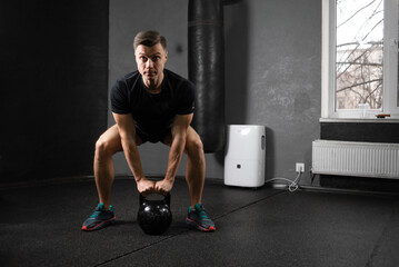 A young male athlete lifts a 20 kg metal kettlebell with two hands from a bent knee position. Training in the gym to raise the metal.