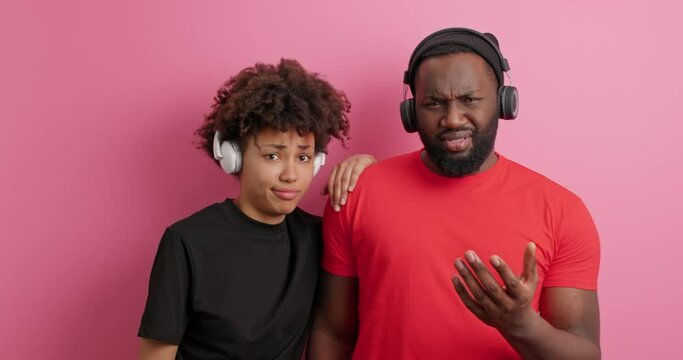 Puzzled displeased ethnic woman and man with dark skin look indignant at each other shrug shoulders listen music viw wireless headphones cannot understand why something happened so pose indoor