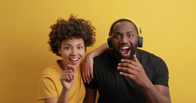 Impressed dark skinned woman and man friends say wow and point directly at camera cannot believe in something notice incredible thing wears yellow black t shirt pose indoor. Reaction concept