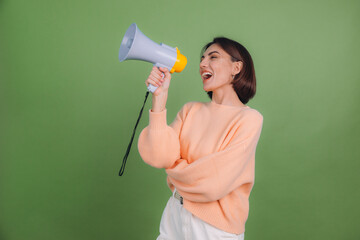 Young woman in casual peach sweater isolated on green olive color background happy funny screaming in megaphone