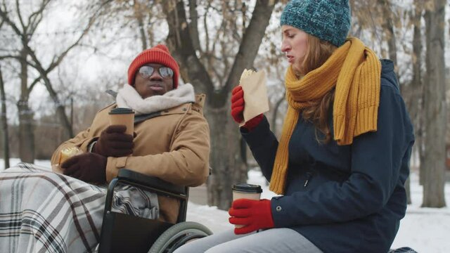 Young Afro-American man on wheelchair and Caucasian woman sitting outdoors on winter day, eating street food, drinking to go coffee, chatting and laughing
