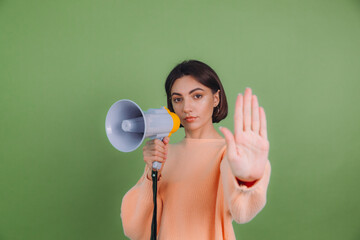 Young woman in casual peach sweater isolated on green olive color background unhappy serious with megaphone doing stop sing with palm of the hand. Warning expression