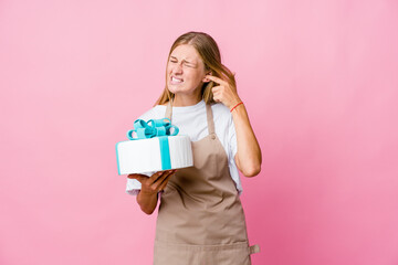 Young russian baker woman holding a delicious cake covering ears with hands.