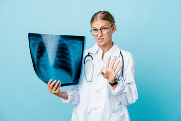 Young russian doctor woman holding a bone scan on blue rejecting someone showing a gesture of...