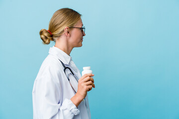 Young russian doctor woman holding pills bottle on blue gazing left, sideways pose.