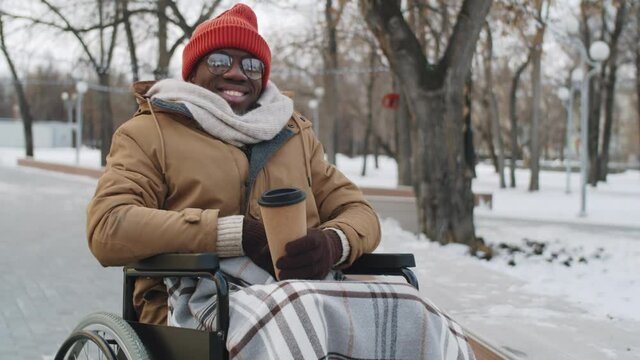 Portrait of young disabled Afro-American man on wheelchair holding disposable coffee cup, looking at camera and smiling while posing in park on winter day