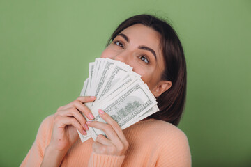 Young woman in casual peach sweater  isolated on green olive background  lucky holding fan of 100 dollar bills funny smell cash money copy space