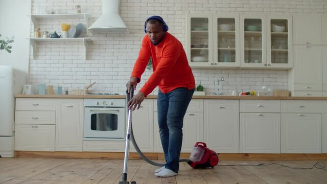 Positive cheerful handsome african man enjoying catching music in wireless headphones, vacuuming kitchen floor and swaying to the beat while making housework during spring cleanup at weekend.
