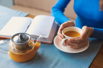 Woman have a teabreak during working hours and planning next work day. Womans hands holding a cup of vitaminic herbal healthy tea from orange and berry sea buckthorn, sitting in cafe alone ang writing
