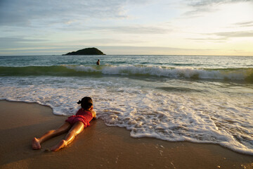 Obraz premium asian child happy or kid girl lie down on stomach hand to chin on island sand beach and sea with family on golden warm sunlight to morning sunrise or sunset for vacation travel on summer holiday relax