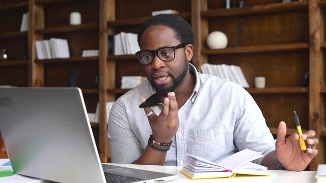 Confident African-American businessman in glasses using voice recognition function on a smartphone. Modern multiracial male entrepreneur sending a vocal message via online messenger, a laptop near