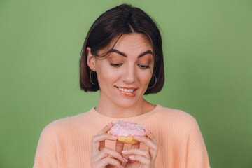 Young stylish woman in casual peach sweater and isolated on green olive background look at pink donut with hunger biting lip copy space