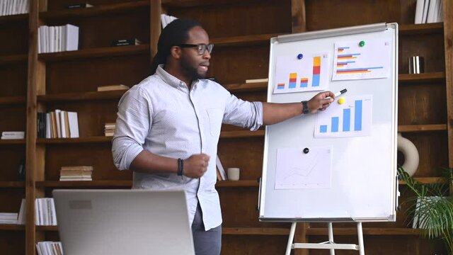 Young African American employee standing explaining project results in graphs, streaming lesson online and looking at webcam, pointing on whiteboard, talking with the virtual group online via laptop